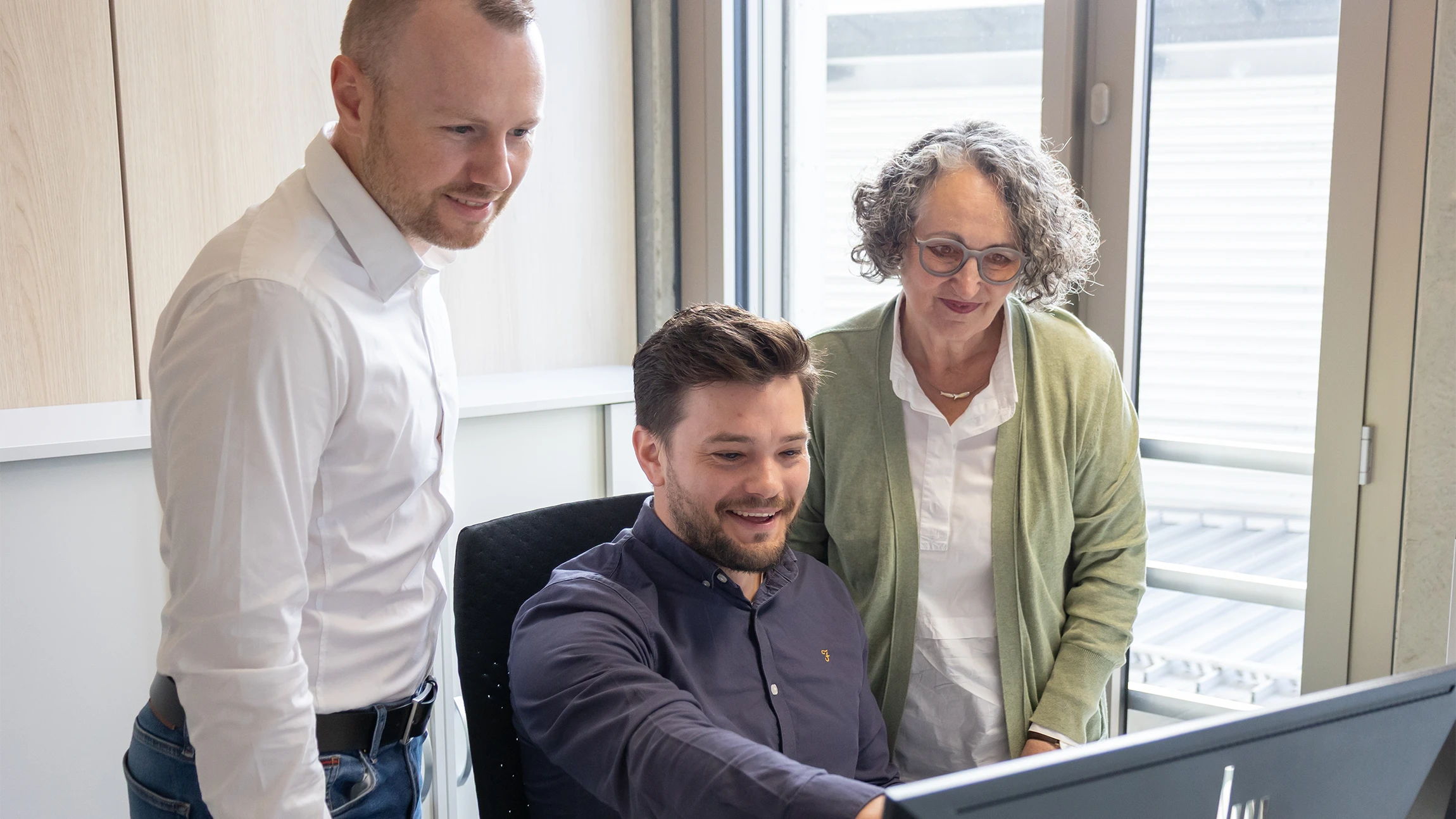Three people standing and sitting around a desk with a computer monitor in an office with large windows.