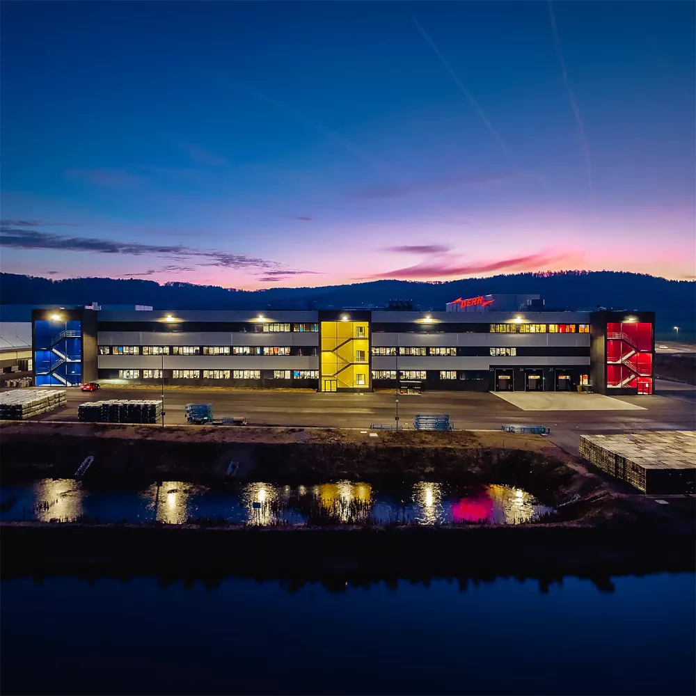 Modern industrial building at dusk with illuminated windows, colorful stairwells, loading bays, and reflections in a water basin.