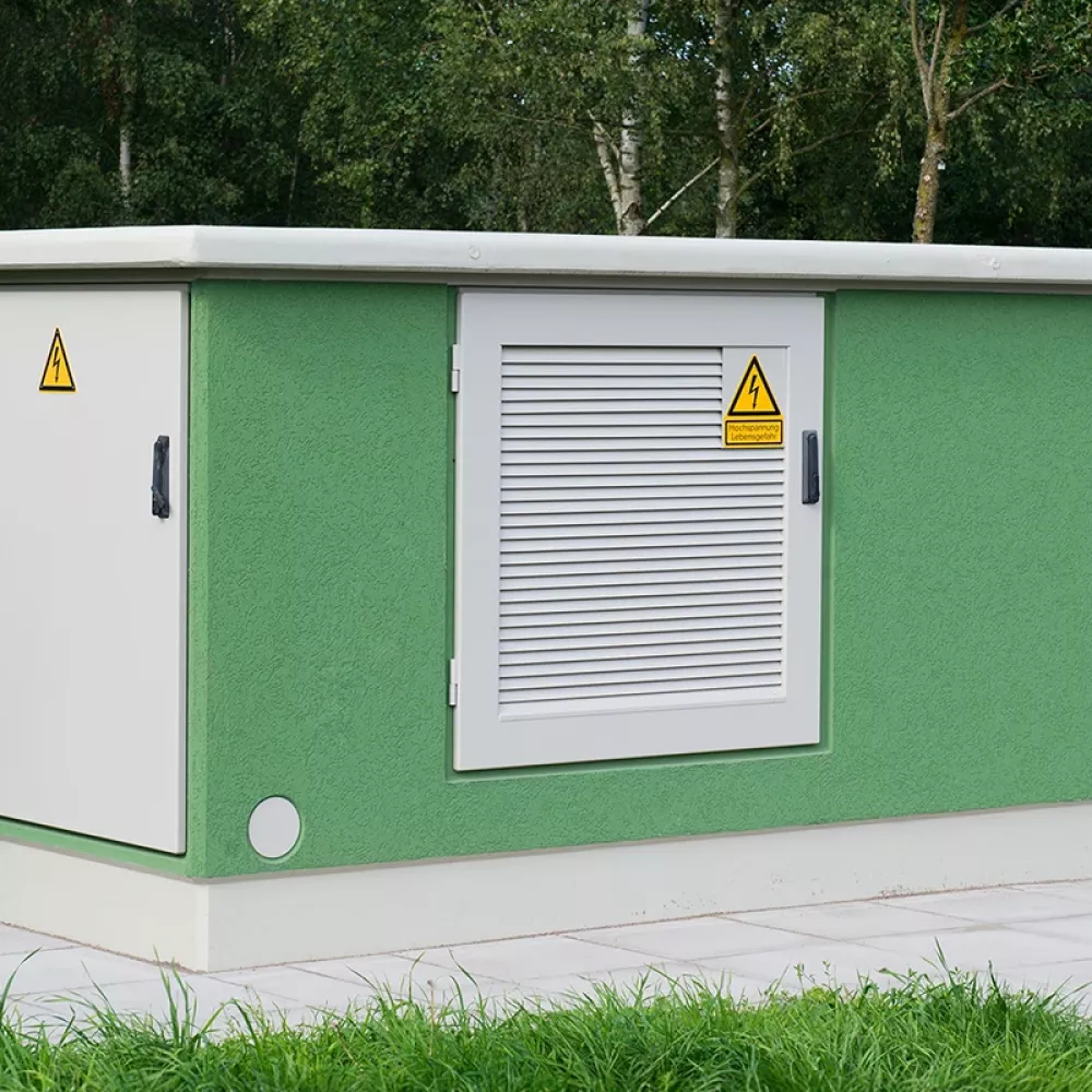 Green transformer station building with ventilation door and high-voltage warning signs, surrounded by grass and trees.