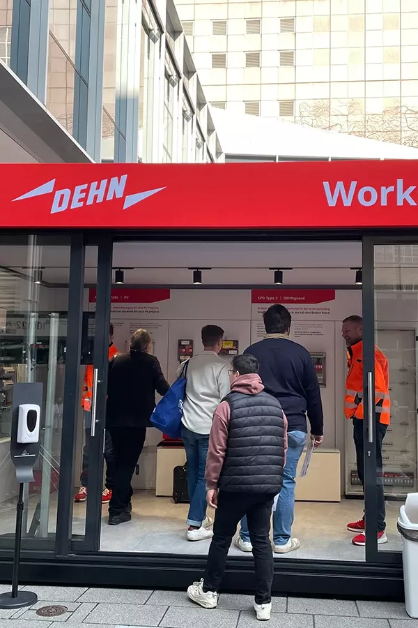 An outdoor display booth with the sign 'DEHN Work' in red letters. People are interacting with staff and viewing materials inside the booth, which has a glass exterior in a modern urban setting.