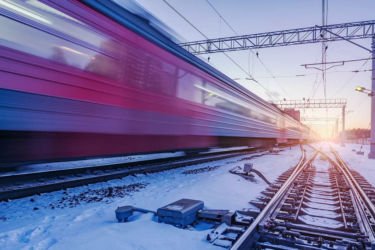 High-speed train passing a snow-covered railway junction at dusk, with blurred motion, overhead power lines, and visible switch components along the tracks.
